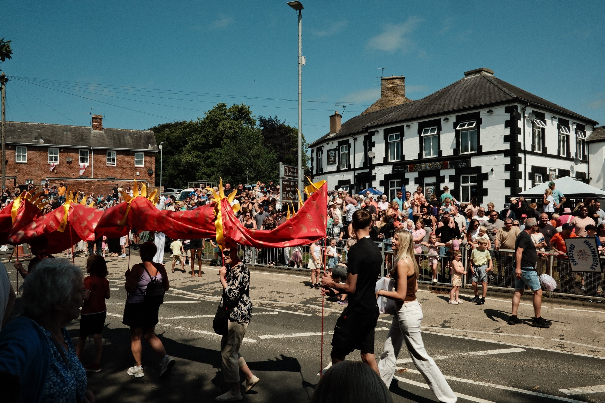 People carrying a dragon costume above their heads pass a crowd of people in front of a white pub.